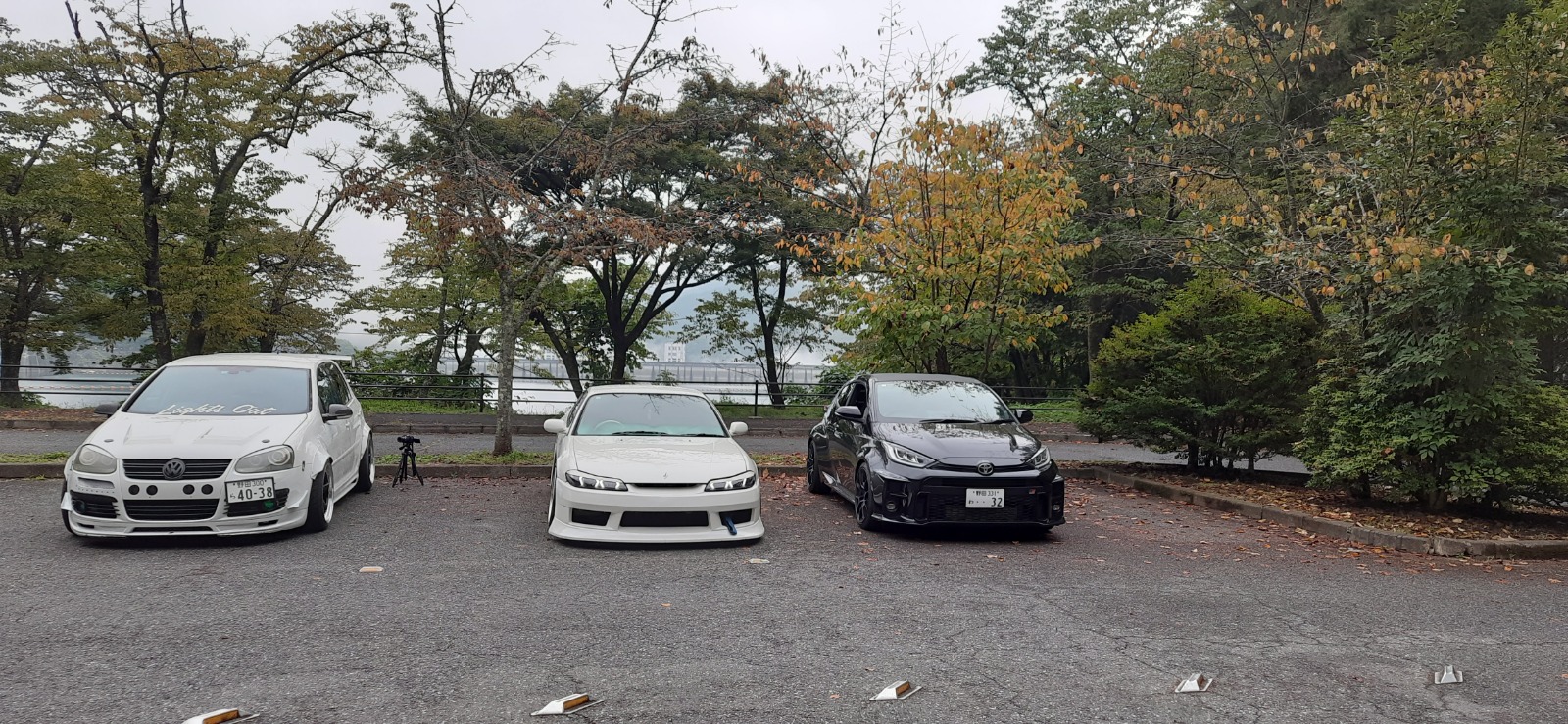 Three modified cars — a VW, a Nissan Silvia, and a black hatchback — parked by a lake with autumn trees on Mount Hinohara