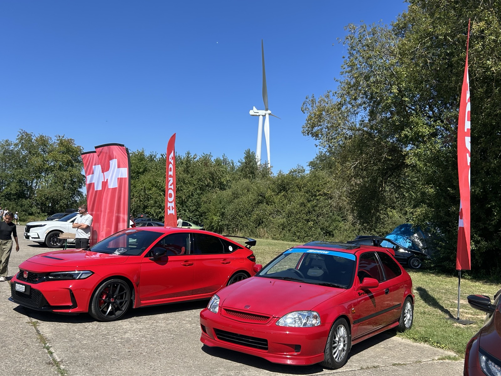 The red Civic EK parked next to a new Honda Civic Type R at a Honda Euro meet, with a wind turbine in the background